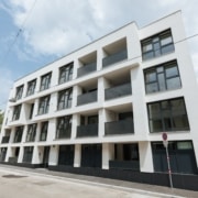 Dreistöckiges Wohnhaus mit heller Fassade und dunkelgrauen Fensterrahmen und Straße die davor vorbeiführt.