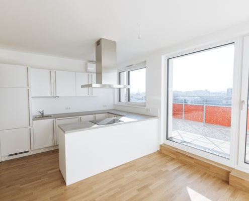 Die Küche hat weiße Schränke, eine Nirosta-Dunstabzugshaube und einen hellen Parkettboden. Der Wohnraum hat große Fenster und führt zur Terrasse.