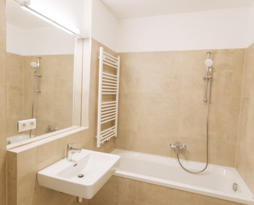Das Bad hat eine Badewanne, ein Waschbecken, einen Spiegel und einen Handtuchtrockner. Die Fliesen sind beige.