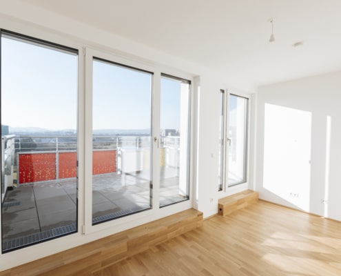 Der Wohnbereich hat große Fenster und einen hellen Parkettboden. Von hier aus kann man auf die Terrasse schauen.