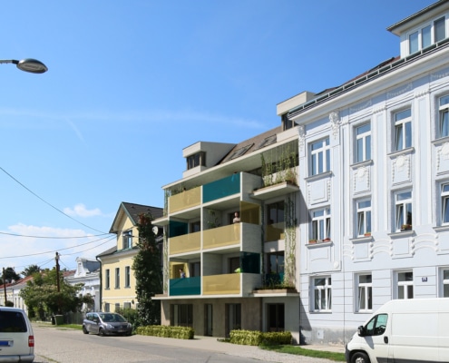Hier sieht man mittig ein Haus mit beigen und petrolfarbenen Balkonen.