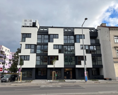 Hier ist ein Haus mit heller Fassade umd einer Straßenlaterne zu sehen. Davor verläuft eine Straße.