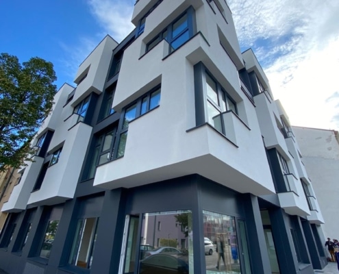 Hier sieht man ein Eckhaus. Im unteren Bereich ist die Fassade dunkelgrau und nach oben weiß mit grauen Fensterrahmen.