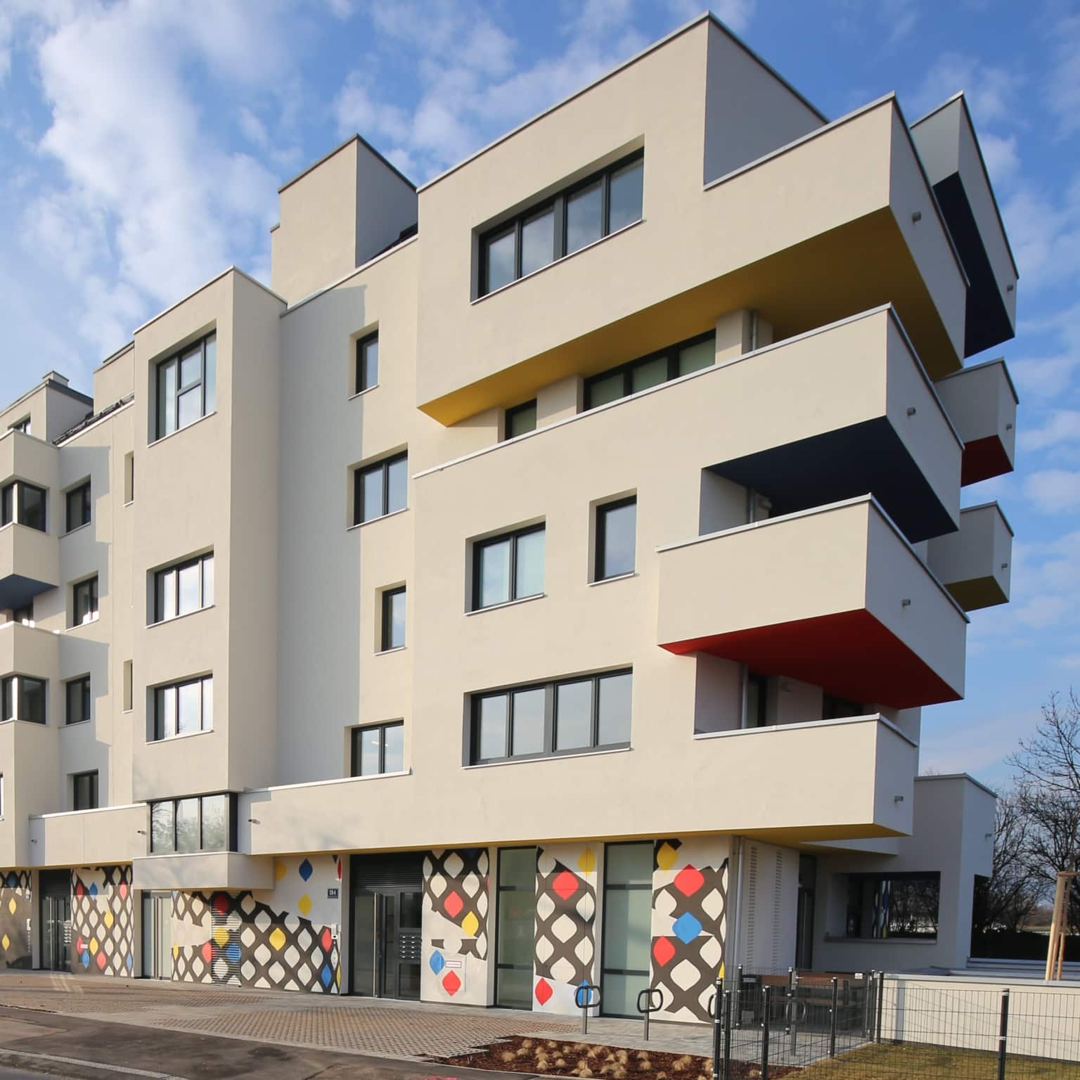 AM-quadratisch Das Bild zeigt einen Teil des Hauses heller Fassade und seitlich sieht man Balkone. Im Eingangsbereich gibt es bunte Elemente.